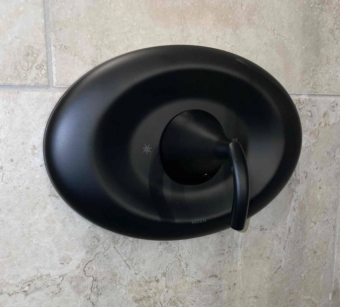 Black Moen single-handle shower control and trim plate against light patterned tile.