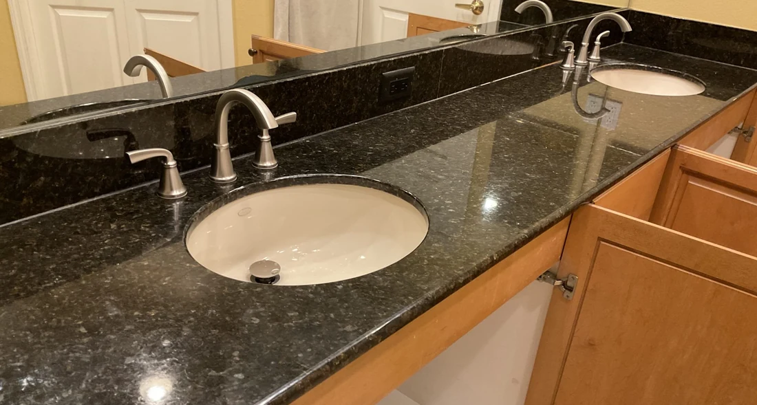 Two brushed-metal faucets with oval inset sinks in a dark vanity countertop.