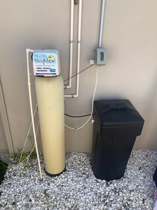 Existing exterior water softener with a tall resin tank, black brine container and connected piping.