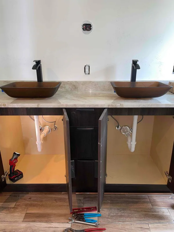 Two vessel sinks with black faucets and exposed under-sink drain connections during vanity work