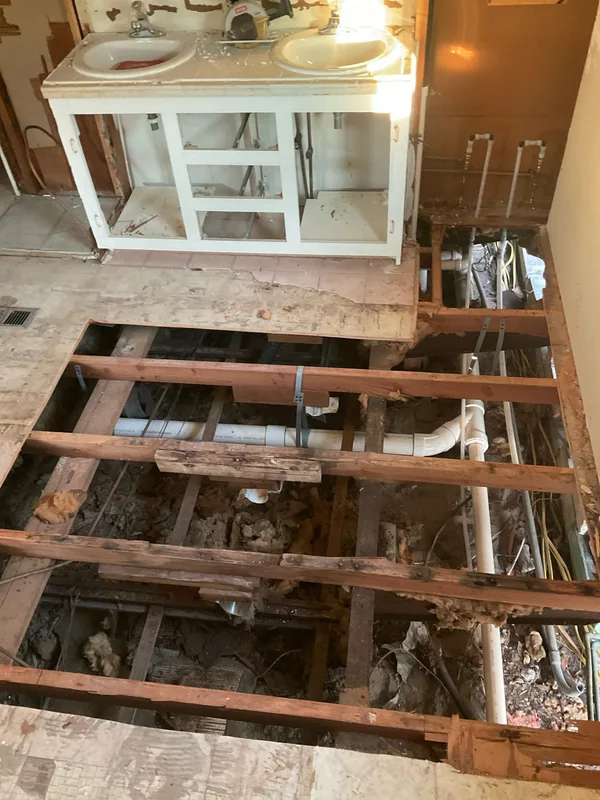 White drain piping and water lines visible beneath open bathroom floor framing beside a vanity
