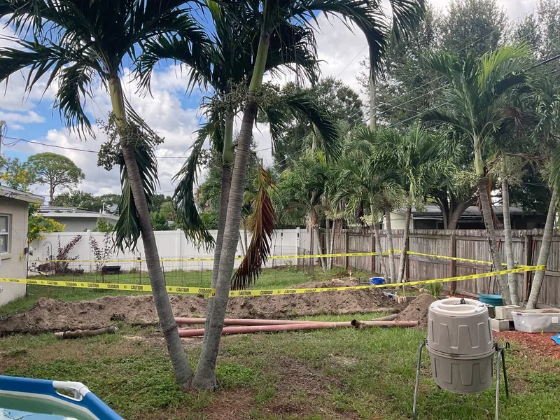 Backyard excavation and pipe sections behind yellow caution tape near palm trees