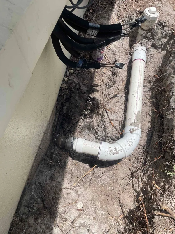 White PVC drain piping in an excavated trench beside an exterior wall beneath insulated service lines