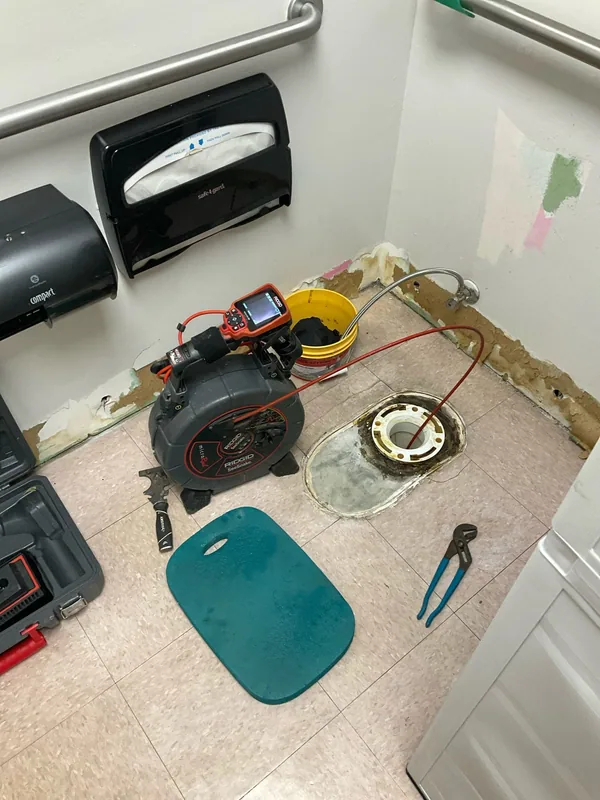 Drain camera reel and monitor beside an exposed toilet flange in a restroom