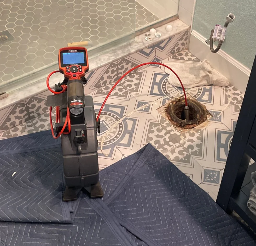 Inspection camera and red cable entering an exposed toilet drain opening beside a tiled shower, with protective floor blankets