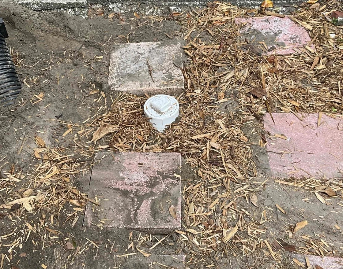 White sewer cleanout cap accessible above backfilled ground beside a walkway and landscape pavers.