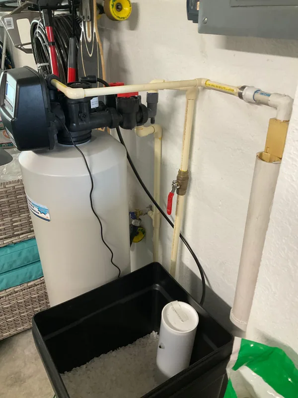 Red-handled brass shutoff valve and connected piping beside a water softener and open brine container.