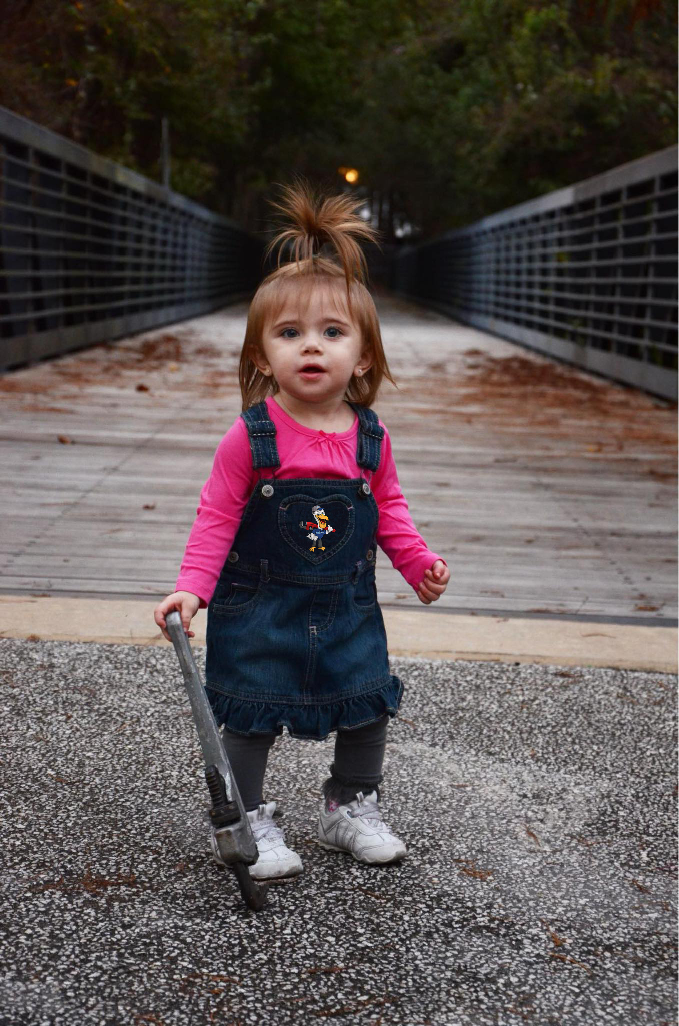 Young girl in pink and denim with a Pelican Ray patch, holding a pipe wrench on a wooden bridge
