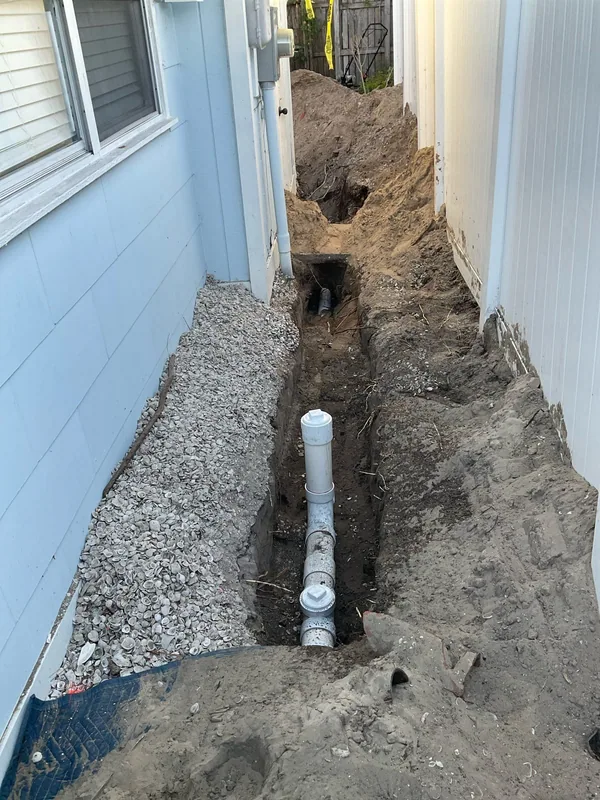 Exposed sewer piping and capped cleanout in a trench beside an exterior wall